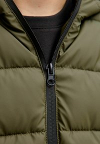 Giacca puffer verde oliva con zip nera, caratterizzata da un design trapuntato e una texture liscia. Fodera interna nera visibile al collo.