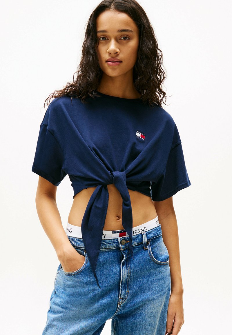 Marineblauwe cropped t-shirt met een frontknoop, korte mouwen en een klein logo-patch, gecombineerd met lichtblauwe high-waisted jeans en een logo-bilband.