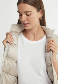 Beige Steppweste mit einem gerippten, hellen Kragen, getragen über einem weißen T-Shirt. Die Weste hat gesteppten Bereiche und einen Reißverschluss.