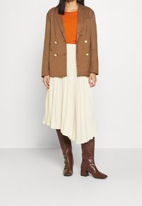 Braun karierter, doppelt geknöpfter Blazer, oranges Oberteil, cremefarbene plissierte Rock und braune, knielange Stiefel aus Leder mit Krokodilstruktur und Blockabsätzen.