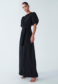 Zwarte maxi-jurk met pofmouwen, een taille met ceintuur en een vloeiende silhouet. Soepele stof, minimalistisch ontwerp, gecombineerd met gouden sandalen met bandjes.