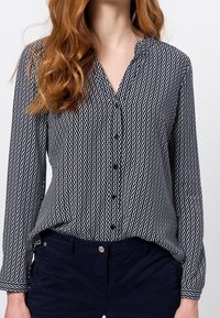 Blusa a righe in bianco e nero con colletto, chiusura frontale a bottoni, maniche lunghe e orlo curvo. Tessuto testurizzato con design ondulato.