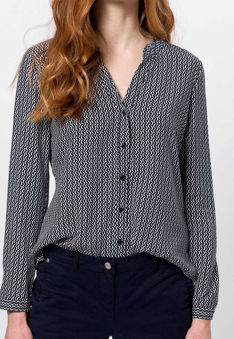 Blusa a righe in bianco e nero con colletto, chiusura frontale a bottoni, maniche lunghe e orlo curvo. Tessuto testurizzato con design ondulato.