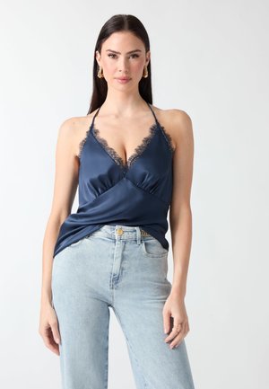 Femme portant un caraco en satin bleu marine avec bordure en dentelle, jean large bleu clair et sandales beiges à talons hauts avec des accents dorés.
