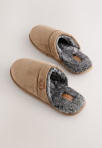 Beige Stoffhausschuhe mit strukturierter Oberfläche und weichem grauem Kunstpelzfutter. Sie verfügen über eine solide, tanfarbene Sohle und einen kleinen Logotag an der Seite.