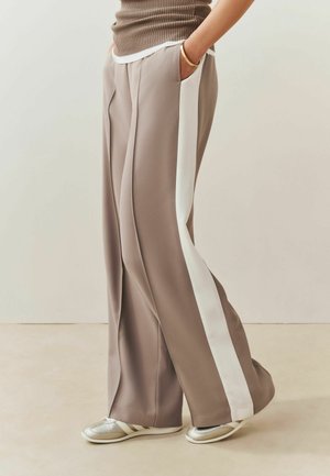 Person trägt weite taupefarbene Hosen mit weißem Seitenstreifen, beige ärmelloses Oberteil, goldenes Armband und silberne Sneakers, steht mit der Hand in der Tasche.