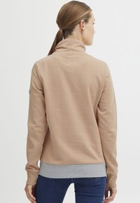 Pullover beige con colletto alto e polsini grigio chiaro. Tessuto morbido con una texture liscia, caratterizzato da un piccolo logo sul collo.