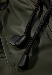 Primer plano de la tela de una chaqueta waterproof verde que muestra cordones negros con extremos texturizados y un botón a presión redondo negro, destacando los detalles de la costura.