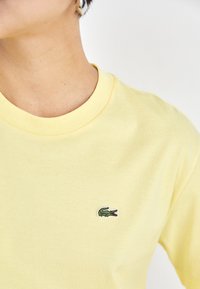 Camiseta de algodón amarilla con cuello redondo, que presenta un pequeño logo bordado de cocodrilo verde en el lado izquierdo del pecho. Textura suave.