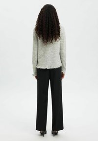 Osoba o długich, kręconych włosach stojąca tyłem, ubrana w jasnoszary sweter i czarne szerokie spodnie z wysokimi obcasami.