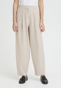 Woman wearing beige wide-leg trousers and black leather loafers, standing against a plain white background.