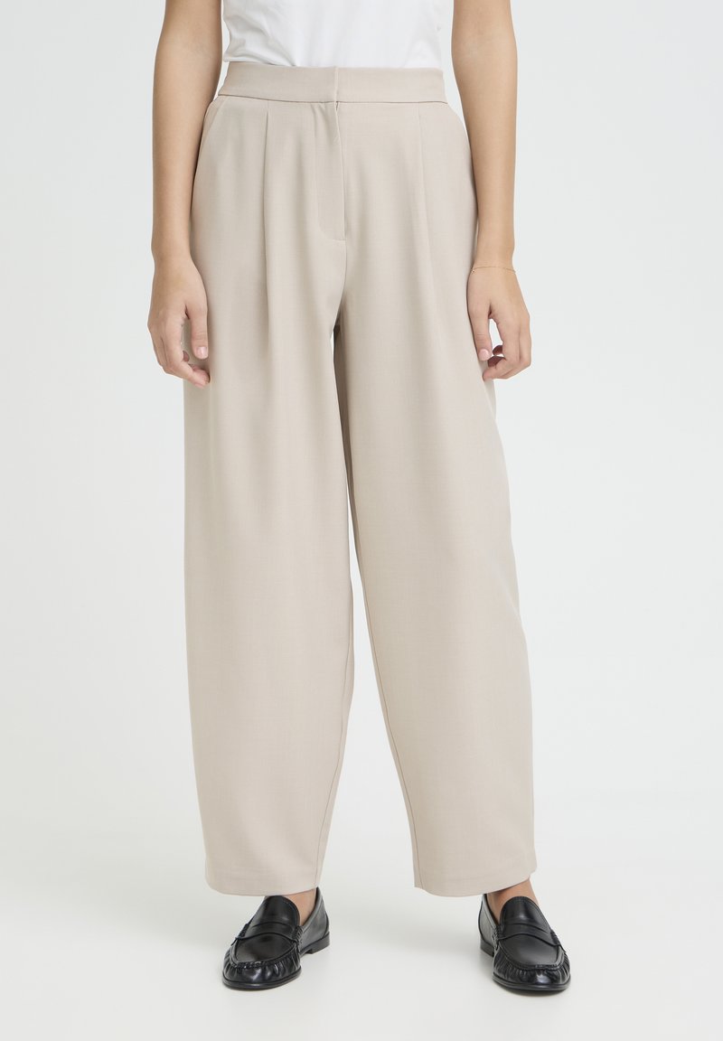 Woman wearing beige wide-leg trousers and black leather loafers, standing against a plain white background.