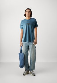 Camiseta de algodón azul con mangas cortas, que presenta un estampado gráfico. Combinada con jeans de mezclilla azul claro y una bolsa de tela azul.