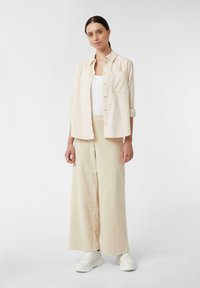 Camicia oversize beige con tasca, abbinata a pantaloni beige a gamba larga. Tessuto morbido, indossata con sneakers bianche. Palette di colori semplice e neutra.