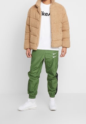 Tan fleece-dunjakke med vandret syning, hvid t-shirt med sort logo, grønne træningsbukser med sorte sideaccenter og hvide sneakers.