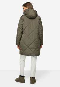 Cappotto invernale trapuntato verde oliva con cappuccio, con un motivo a diamante e maniche lunghe, indossato sopra pantaloni chiari e stivali.