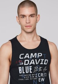 Črn bombažni top brez rokavov z grafičnim dizajnom, ki vključuje mešanico besedila v beli, modri in rdeči barvi, okroglo ovratnico.
