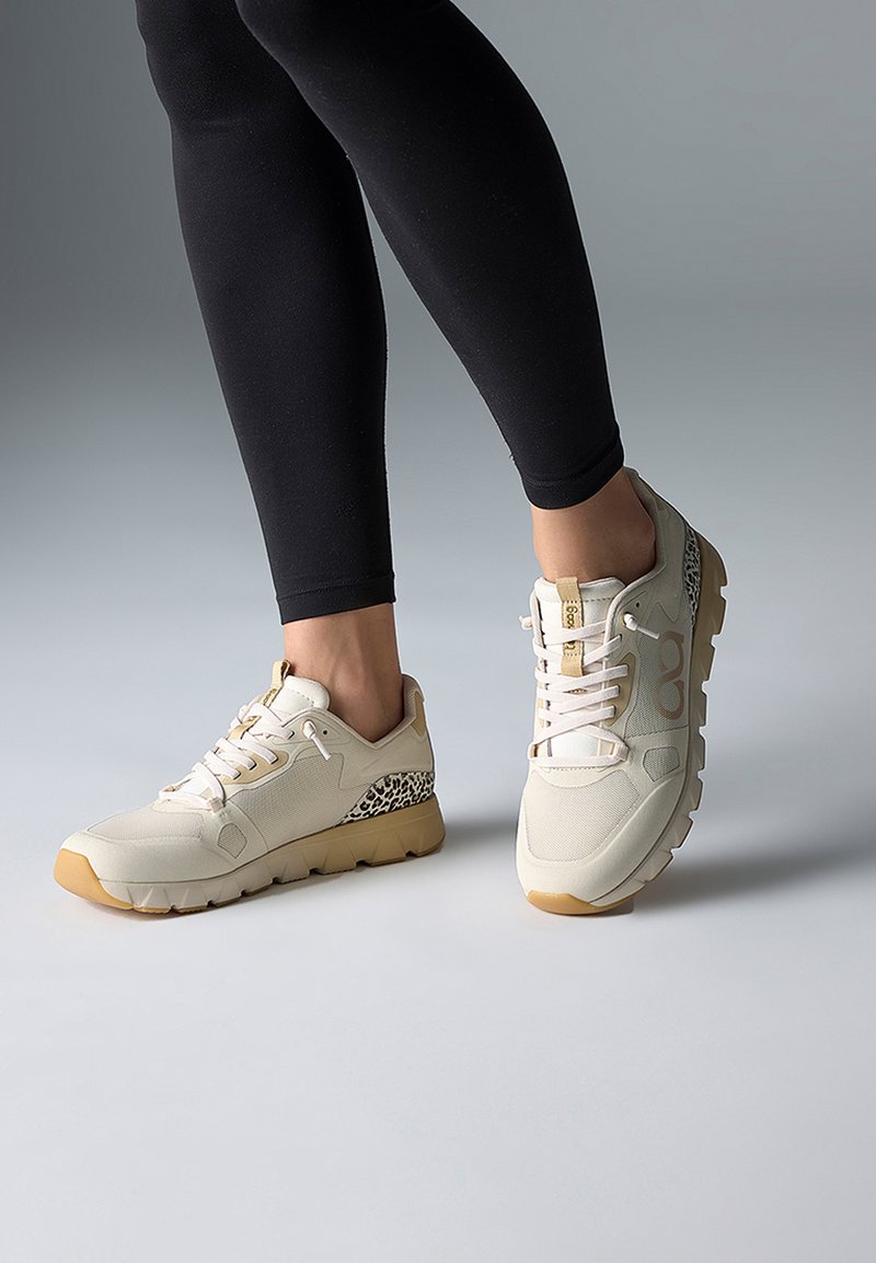 Ben i sorte leggings iført beige sneakers med hvide snørebånd og leopardmønstrede detaljer, stående på en grå overflade.