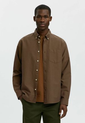 Homme faisant face vers l'avant, portant une chemise marron déboutonnée sur un t-shirt marron et un pantalon vert foncé, mains partiellement dans les poches, expression neutre.
