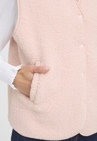 Pasztell rózsaszín bolyhos mellény, textúrált felülettel, gombos záródással és oldalsó zsebekkel. Az anyag puha, kényelmes megjelenésű.