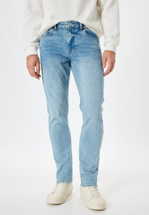 Jeans de mezclilla azul claro con corte ajustado, que presentan cinco bolsillos, pasadores para el cinturón y un ligero desgastado, combinados con zapatillas de color crema.