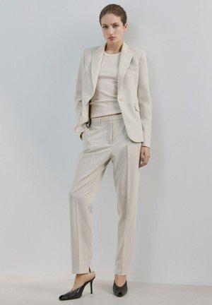 Kvinde står op ad en hvid væg iført et beige jakkesæt med matchende bukser, blazer, cremefarvet ribbet top og sorte højhælede sko.