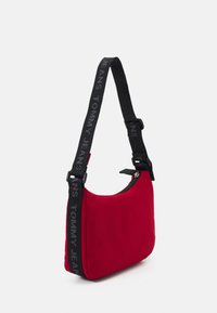 Bolso de hombro de tela roja con forma curva, banda negra con la marca "TOMMY JEANS" y cierre superior con cremallera. Diseño sencillo.