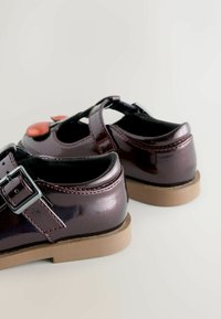 Dunkelburgunderfarbene Lacklederschuhe mit einem vorderen Riemen, silberner Schnalle und braunen Gummisohlen, versehen mit rotem Stich und einem runden Akzent.