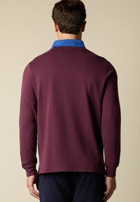 Długosłodkowy polo w głębokim burgundzie z kontrastowym niebieskim kołnierzem. Miękka, gładka tekstura z luźnym krojem i detalami szwów na plecach.