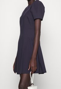 Marineblaues Kurzarmkleid mit tailliertem Oberteil und ausgestellt Rock. Verfügt über seitliche Taschen und einen glatten, strukturierten Stoff. Weißes Handtaschenaccessoire.