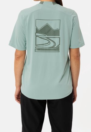 Person iført en lysegrøn kortærmet skjorte med sort bjerg- og vejgrafik på ryggen, kombineret med sorte bukser.