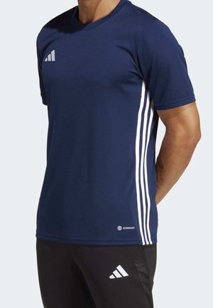Mężczyzna w granatowej koszulce Adidas z białymi paskami po bokach i czarnych spodniach Adidas z logo na lewym udzie, stoi z rękami za plecami.