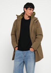 Kaki pufferjakke med hætte, lynlås og trykknapper. Har et logomærke og er iført over en sort sweater og blå jeans.