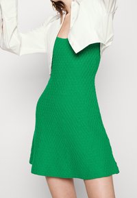 Abito in maglia verde con texture, caratterizzato da un corpino aderente e una gonna svasata. Indossato con una camicia bianca oversize per un effetto sovrapposto.