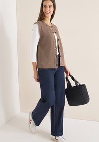 Femme souriante, portant un gilet en maille beige boutonné sur un t-shirt blanc à manches longues, jean bleu foncé, baskets blanches, tenant un sac à main noir, debout devant un mur uni.
