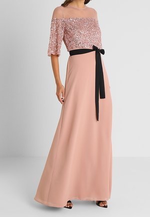 Femme portant une robe rose à manches longues jusqu'au sol avec un top à sequins, un col transparent et une ceinture noire à la taille, debout sur un fond uni.