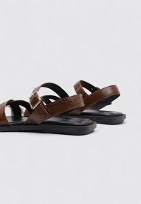 Las sandalias de imitación de cuero marrón presentan una textura de cocodrilo, una correa ajustable con hebilla y una suela negra plana con elementos de diseño minimalistas.