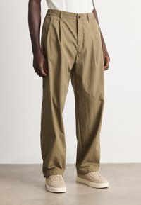 Dark-skinned person wearing loose beige trousers and cream-colored sneakers, standing on a gray floor against a white wall.