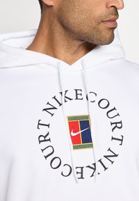 Bela kapuca s vrvicami, ki ima barvit izvezen etiketo in besedilo "NIKE COURT" v krožni zasnovi. Nežen občutek tkanine.