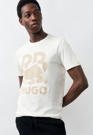 Νεαρός άνδρας που φορά λευκό μπλουζάκι με μπεζ λογότυπο ταύρου και το κείμενο "RB HUGO", στέκεται μπροστά σε απλό φωτεινό φόντο.