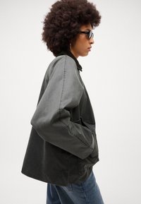 Tamsiai pilkas, per didelis švarkas su struktūrizuotu apykakle, šoninėmis kišenėmis ir matomais siūlių akcentais. Modelis dėvi akinius nuo saulės ir turi garbanotus plaukus.