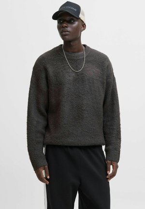 Young man wearing a dark gray fuzzy sweater, black pants, silver chain necklace, and a black and beige cap, standing against a plain white background.