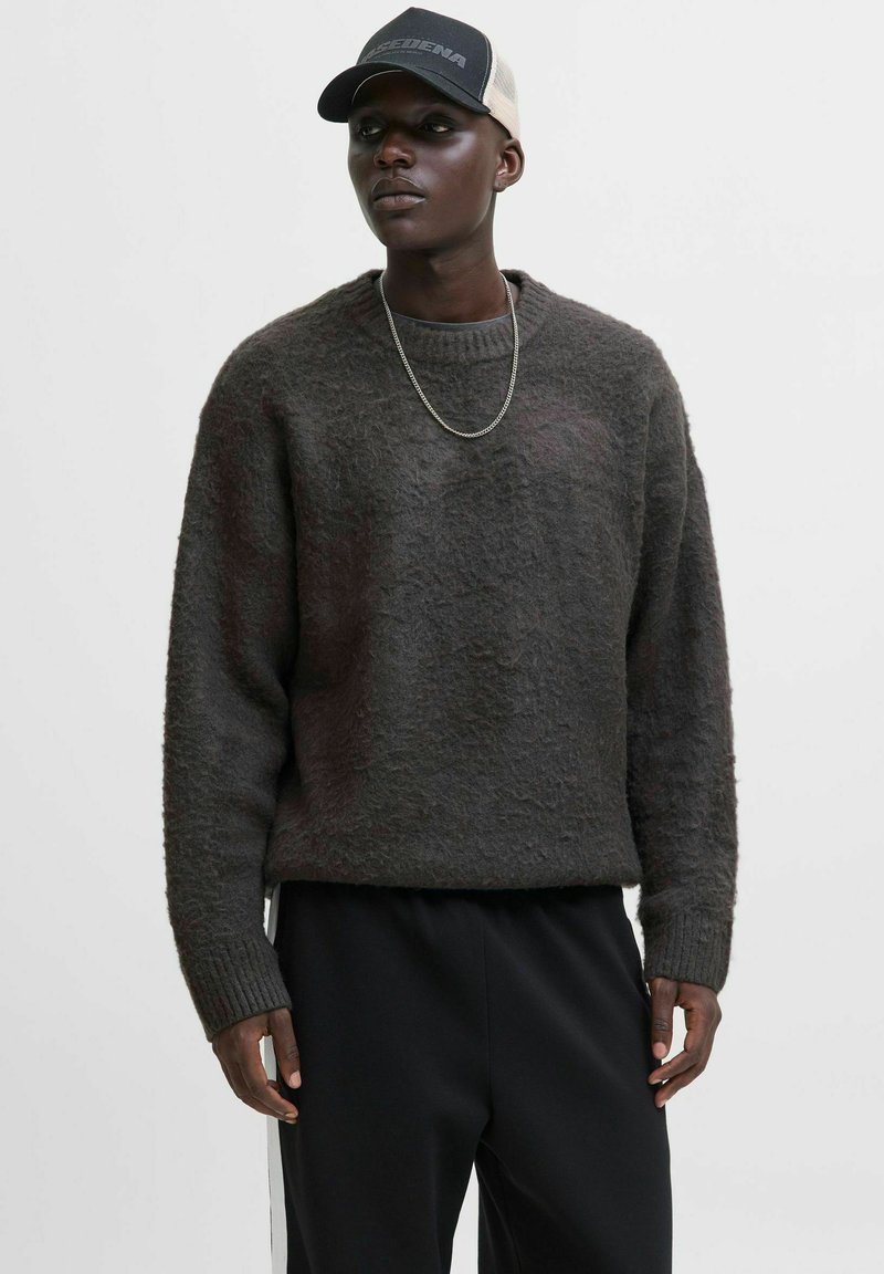 Ung mand iført en mørkegrå, fluffy sweater, sorte bukser, sølv kædehalssmykke og en sort- og beige kasket, der står foran en ensfarvet hvid baggrund.