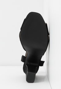 Sandale à talon en suède noir avec un dos ouvert, une large lanière de cheville et une semelle en caoutchouc noir texturée. Doublure intérieure lisse visible.