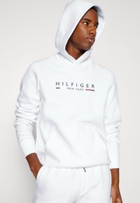 Fehér kapucnis pulóver puha anyagból, elülső zsebbel és húzózsinóros kapucnival. "HILFIGER NEW YORK" felirat színes betűkkel.