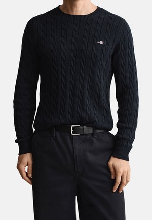 Homme portant un pull noir en maille torsadée avec un petit écusson brodé sur la poitrine, assorti à un pantalon noir et une ceinture noire.