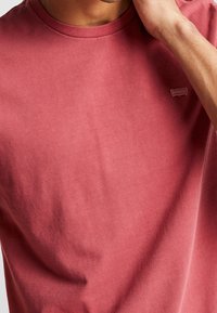 Mann trägt ein verblasstes rotes Rundhals-T-Shirt mit einem kleinen Logo-Patch nahe der linken Schulter und berührt mit seiner rechten Hand sein Gesicht.