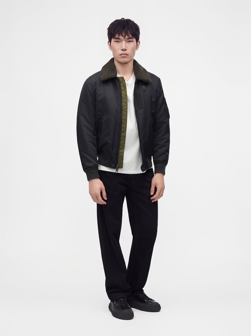 Homme portant une veste bomber noire avec un col vert, un t-shirt blanc, un pantalon noir et des baskets noires, debout devant un fond blanc uni.
