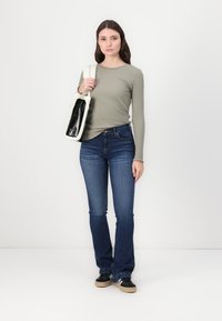 Olive ribbed long-sleeve top, dark blue flared jeans, black and tan sneakers, holding a black and white fur-lined bag. Plain background.