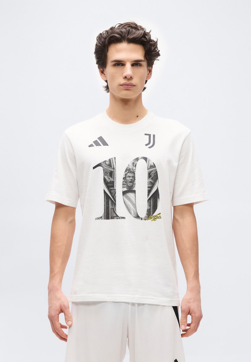 Νεαρός άνδρας που φοράει λευκή φανέλα ποδοσφαίρου της Γιουβέντους με αριθμό 10 και λογότυπα Adidas, στέκεται μπροστά σε μονόχρωμο λευκό φόντο.